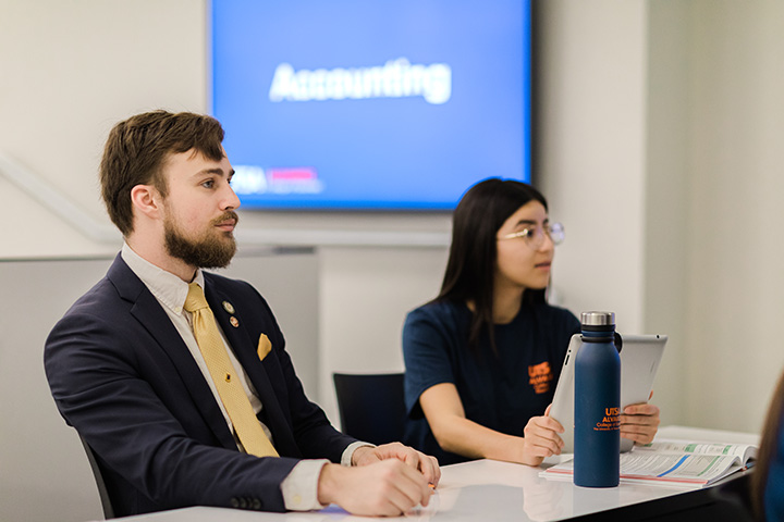 Image of a male student wearing a blue suit and yellow tie and a female student wearing a blue Alvarez College of Business shirt with a screen that says "Accounting" in the background.